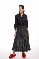 A woman modeling a black long-sleeve button-up shirt and a black pleated midi skirt, paired with red slippers, against a plain white background.