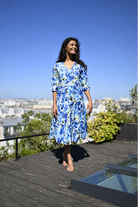 A woman wearing a blue and white floral midi dress walks barefoot on a wooden deck with a city view and clear blue sky in the background.