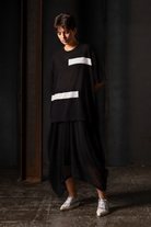 A model wearing a black oversized short-sleeve top with two horizontal white stripe accents and a matching black high-low skirt, paired with white sneakers, standing in a dark industrial setting.