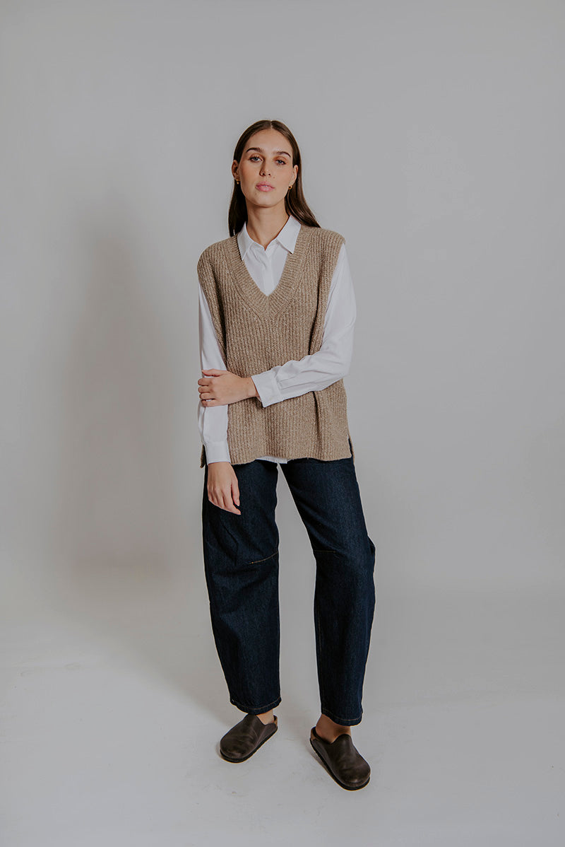 Person wearing a brown vest over a white shirt with dark pants on a plain background