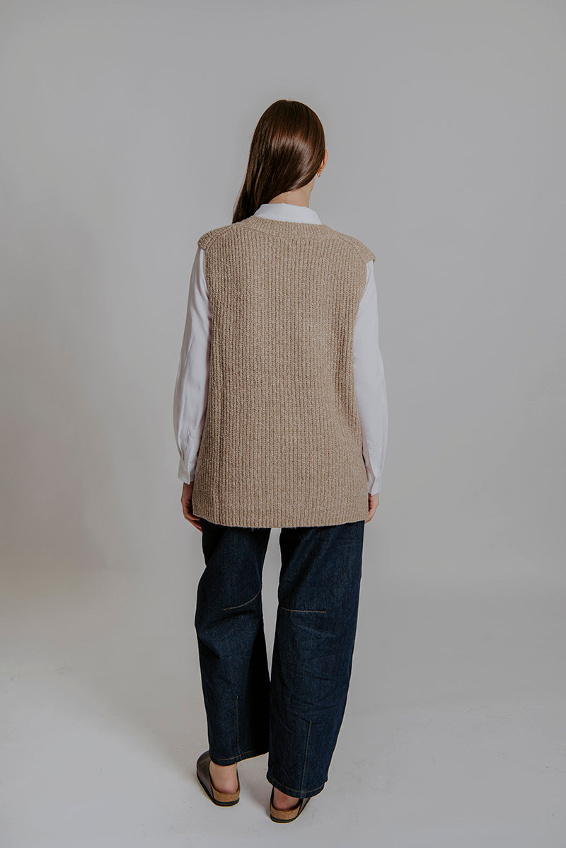 Person wearing a beige vest over a white shirt and dark pants on a plain background