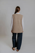 Person wearing a beige vest over a white shirt and dark pants on a plain background