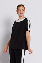A woman modeling a black two-piece set consisting of a short-sleeve top with white shoulder stripes and matching wide-leg trousers with white side stripes, paired with black strappy heels, against a light gray studio background.
