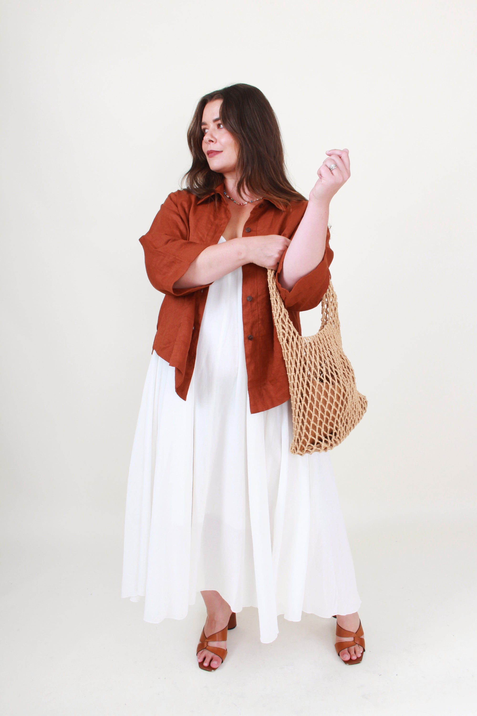 Woman wearing a rust-colored shirt, white dress, and holding a woven bag on a white background