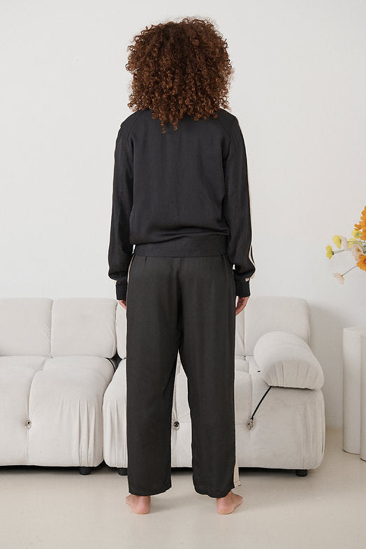 A person standing with their back to the camera in a minimalist room, wearing a black long-sleeve top and black wide-leg trousers with white side stripes. 