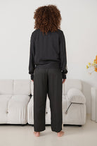 A person standing with their back to the camera in a minimalist room, wearing a black long-sleeve top and black wide-leg trousers with white side stripes. 