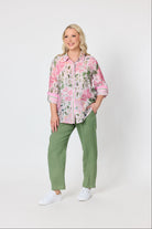 A woman modeling a pink and green floral button-up shirt with rolled sleeves, paired with olive green pants and white sneakers, against a plain light gray studio background.