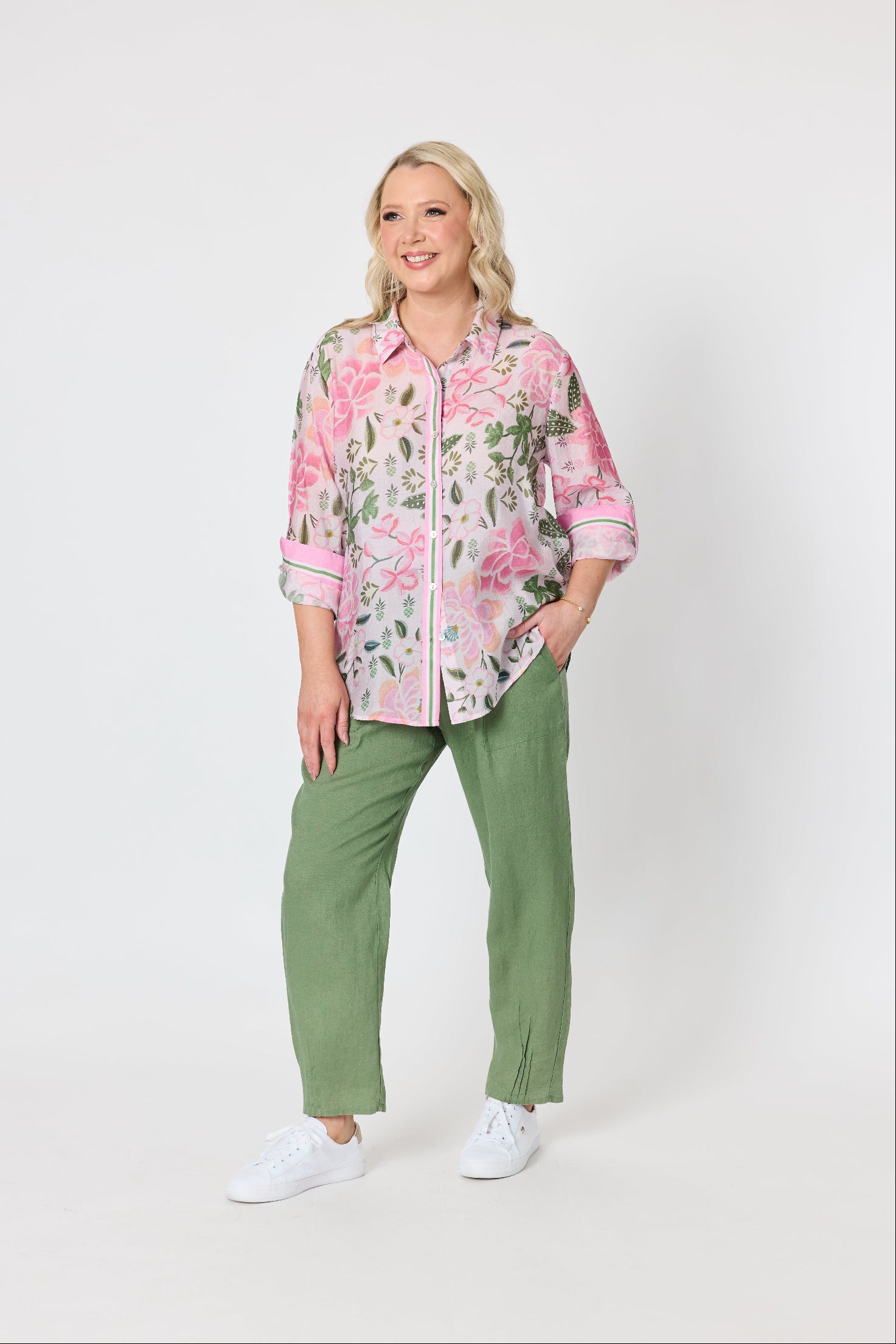 A woman modeling a pink and green floral button-up shirt with rolled sleeves, paired with olive green pants and white sneakers, against a plain light gray studio background.