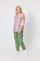A woman modeling a pink and green floral button-up shirt with olive green pants and white sneakers, standing against a plain light gray studio background.