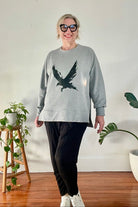 Person wearing a gray sweatshirt with an eagle design, standing indoors with plants in the background.