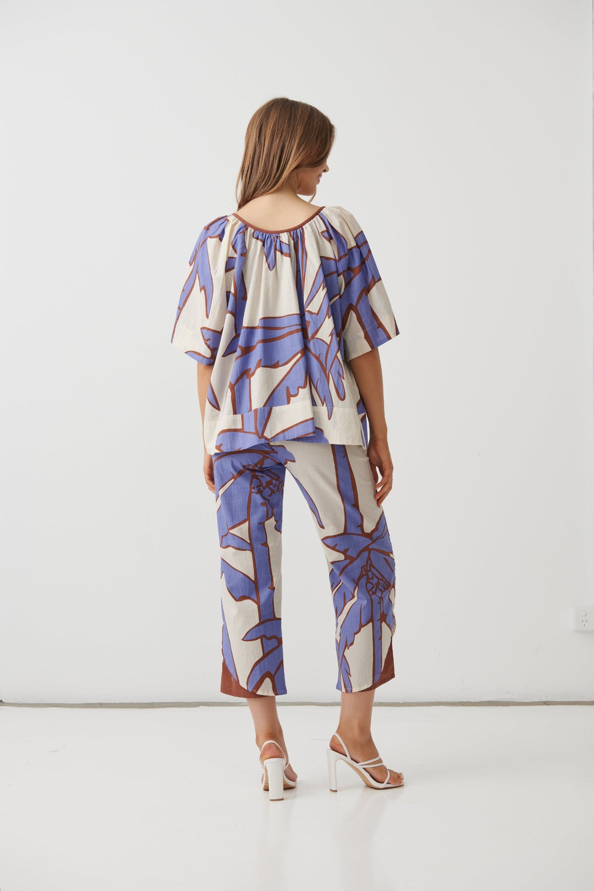 A woman viewed from the back wearing a matching two-piece set with a blue, purple, and brown abstract botanical pattern on a cream background, paired with white high-heeled sandals, set against a plain white studio background.