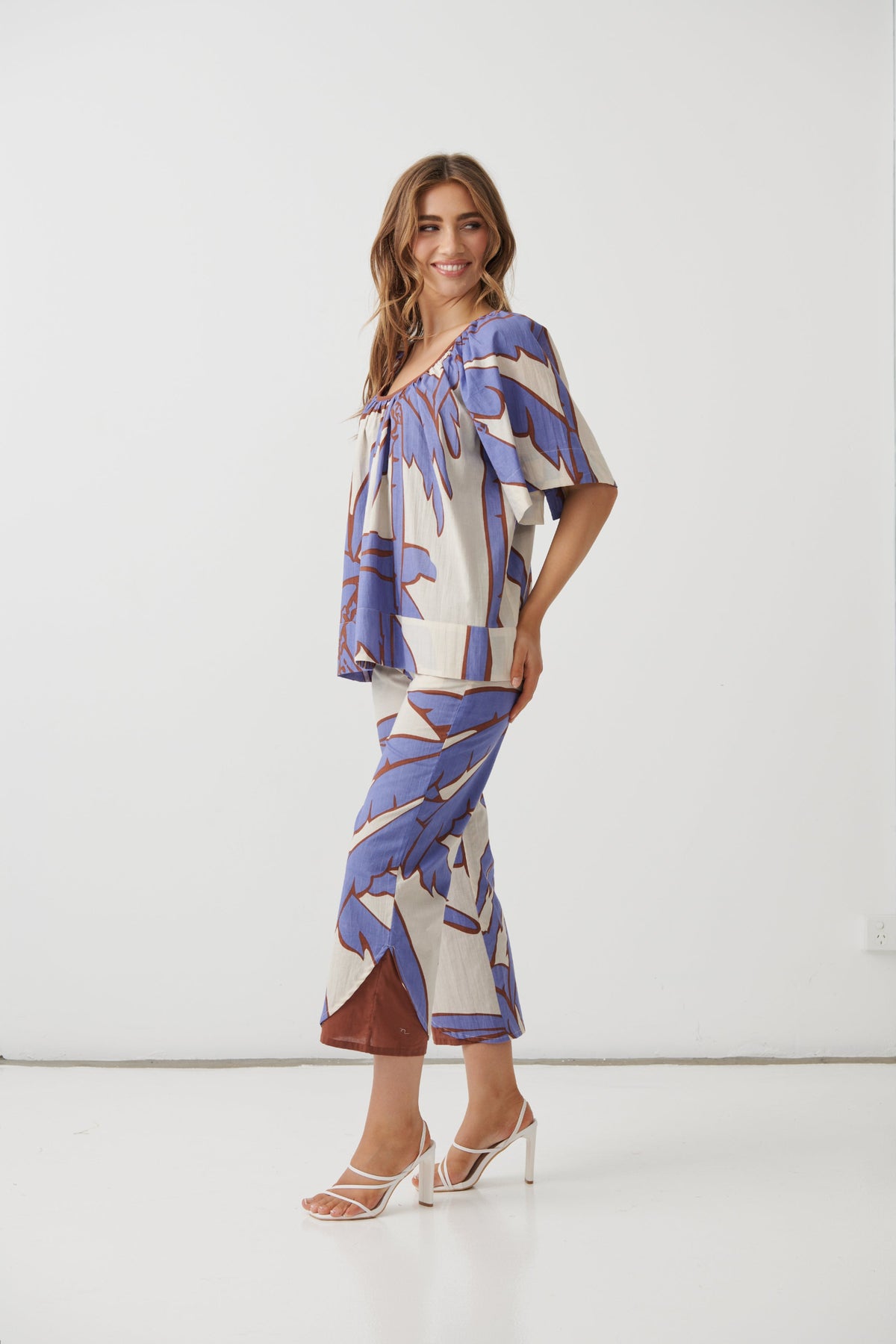 A woman modeling a matching two-piece set featuring a cream base with a blue and brown abstract tropical leaf print, styled with white strappy high-heeled sandals against a plain white studio background.