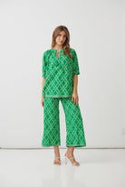 A woman modeling a green and white geometric patterned two-piece set consisting of a short-sleeve top with a keyhole neckline and matching wide-leg cropped pants, photographed against a plain white studio background.