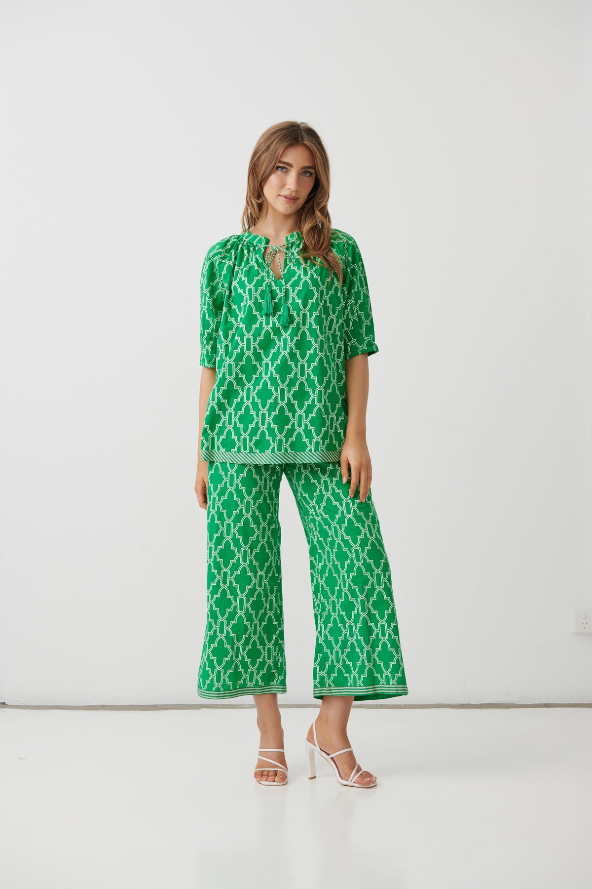A woman modeling a green and white geometric patterned two-piece set consisting of a short-sleeve top with a keyhole neckline and matching wide-leg cropped pants, photographed against a plain white studio background.