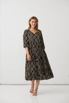 A woman modeling a black midi dress with a beige geometric pattern, standing against a plain white studio background.