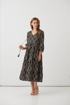 A woman modeling a black midi wrap dress with a beige and brown snake print pattern, holding a black tassel accessory against a plain white studio background.