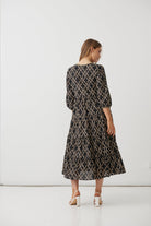 A woman is shown from behind wearing a long-sleeved midi dress with a black and beige geometric trellis pattern, paired with white high heels, set against a plain white studio background.