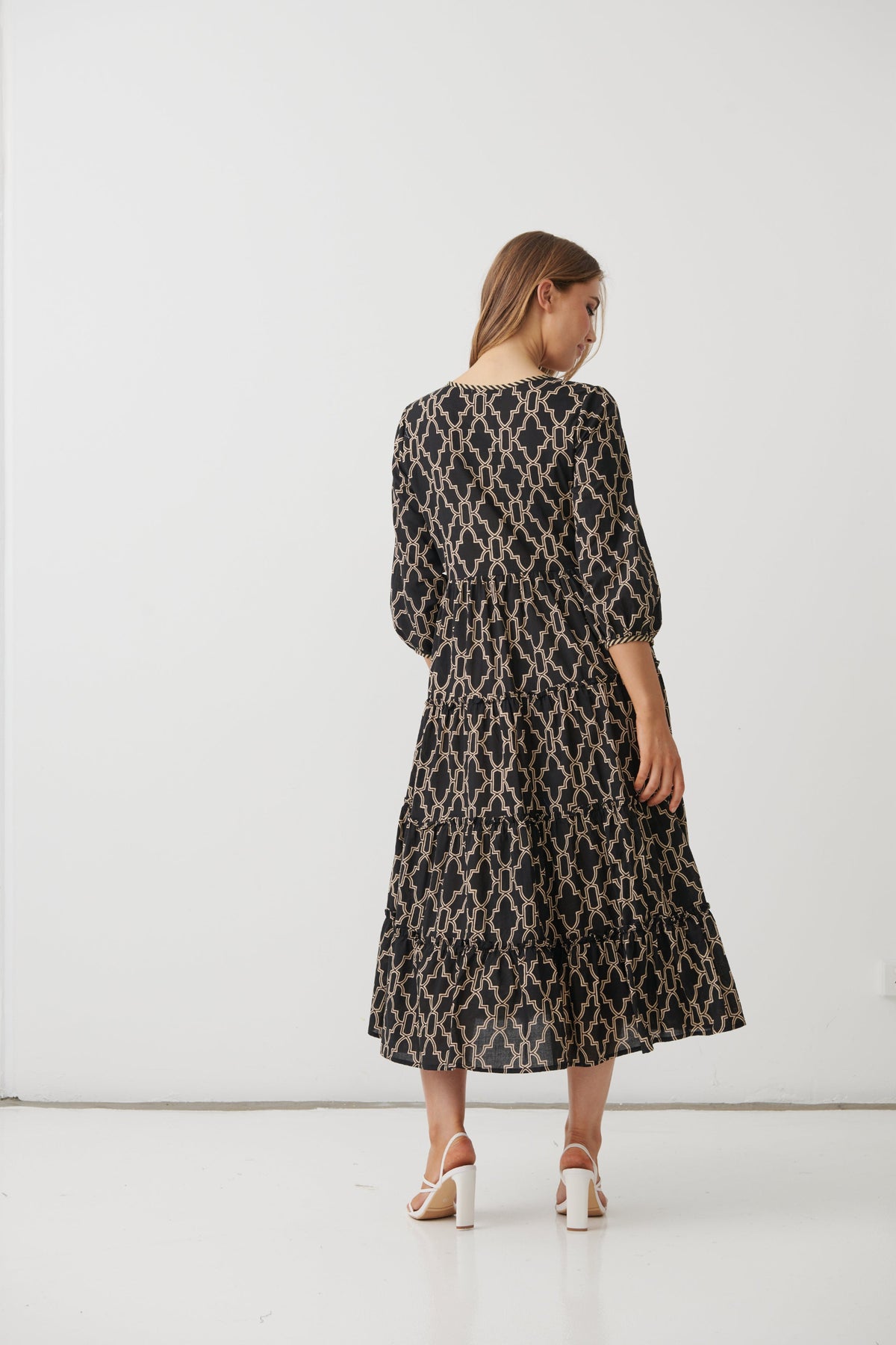 A woman is shown from behind wearing a long-sleeved midi dress with a black and beige geometric trellis pattern, paired with white high heels, set against a plain white studio background.