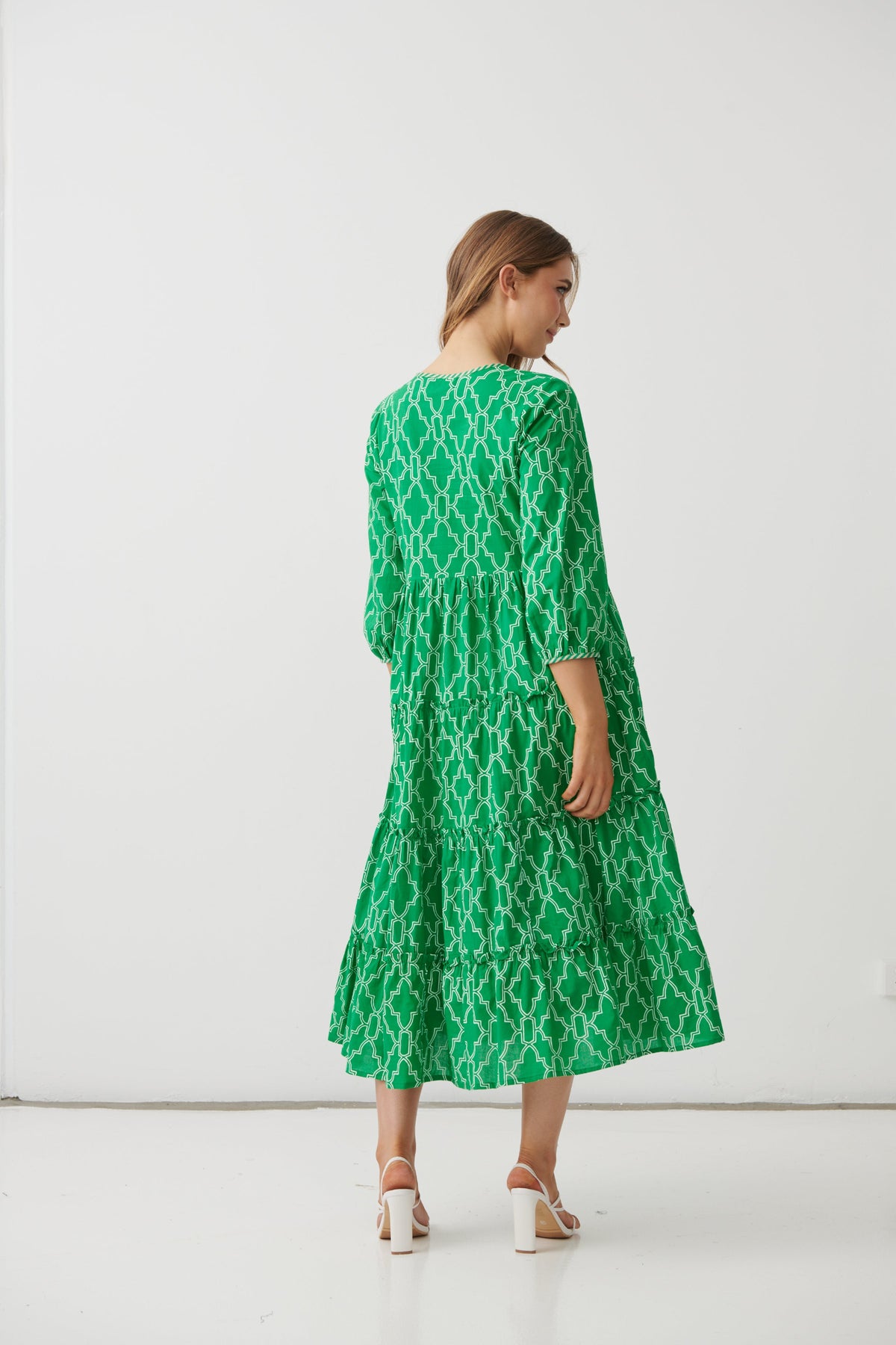 A woman wearing a green midi dress with a white geometric trellis pattern, shown from the back in a studio setting with a white background.