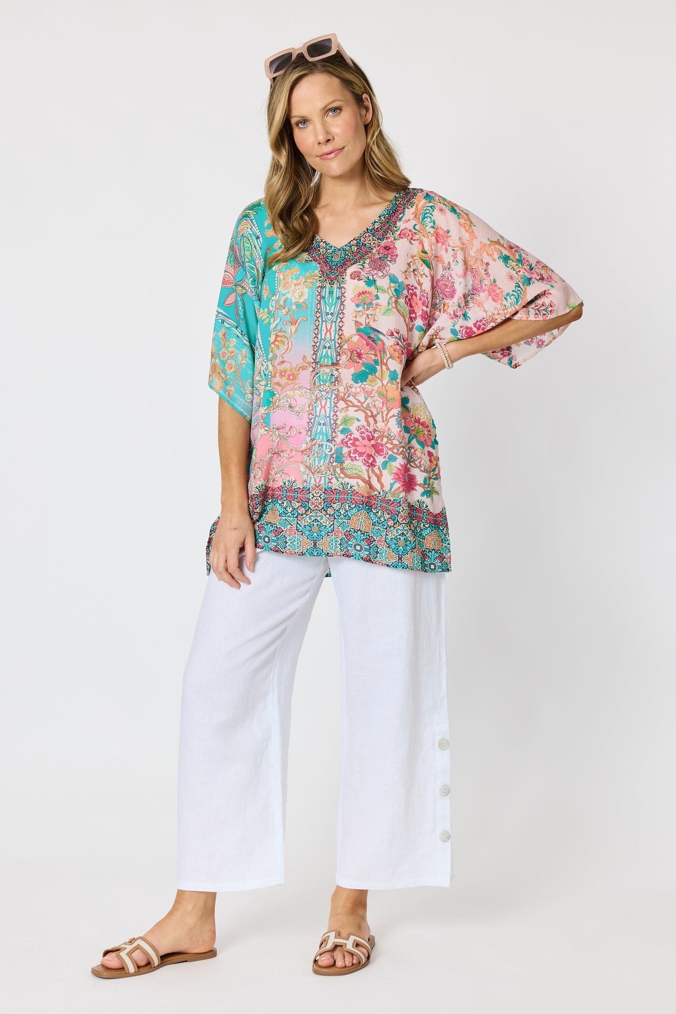 Woman wearing a colourful pink and blue printed floaty top with elbow length sleeves and detailed border and neckline and white pants against a white background.