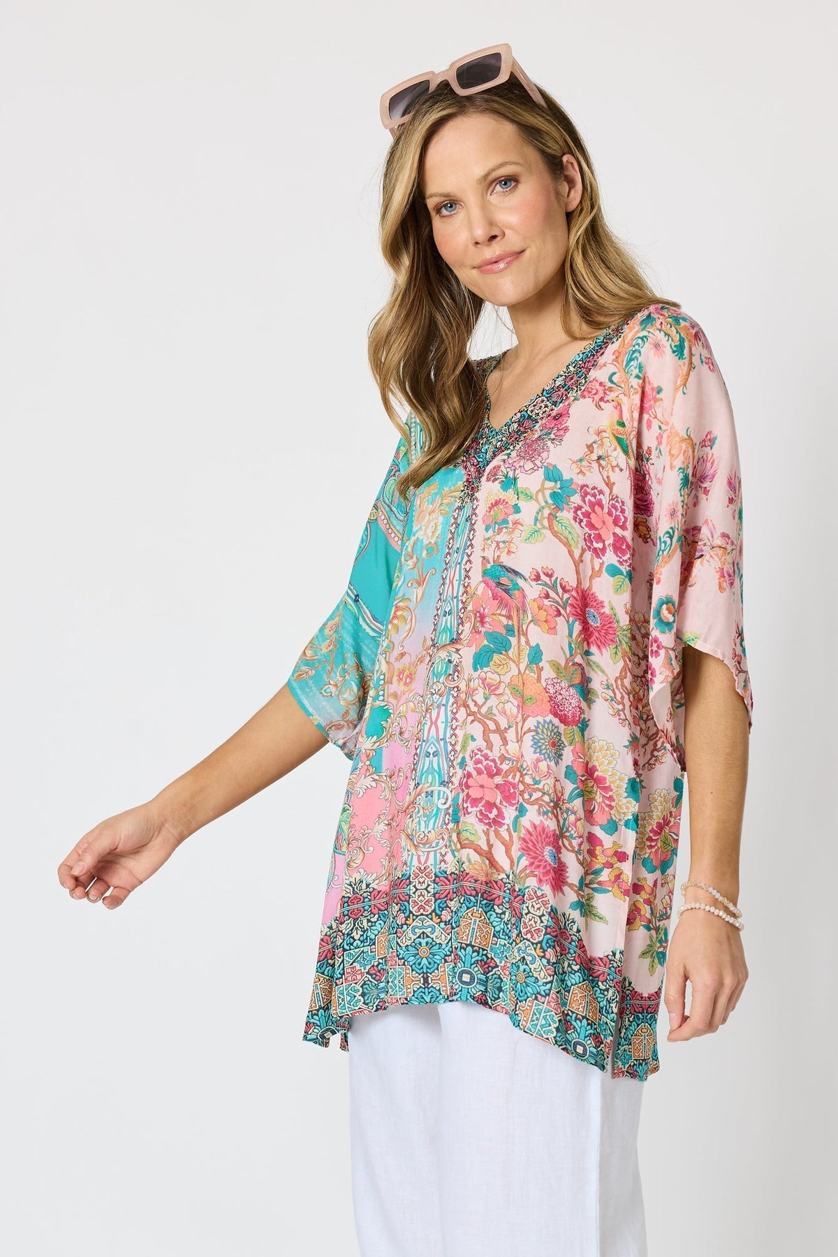 Woman wearing a colourful pink and blue printed floaty top with elbow length sleeves and detailed border and neckline and white pants against a white background.