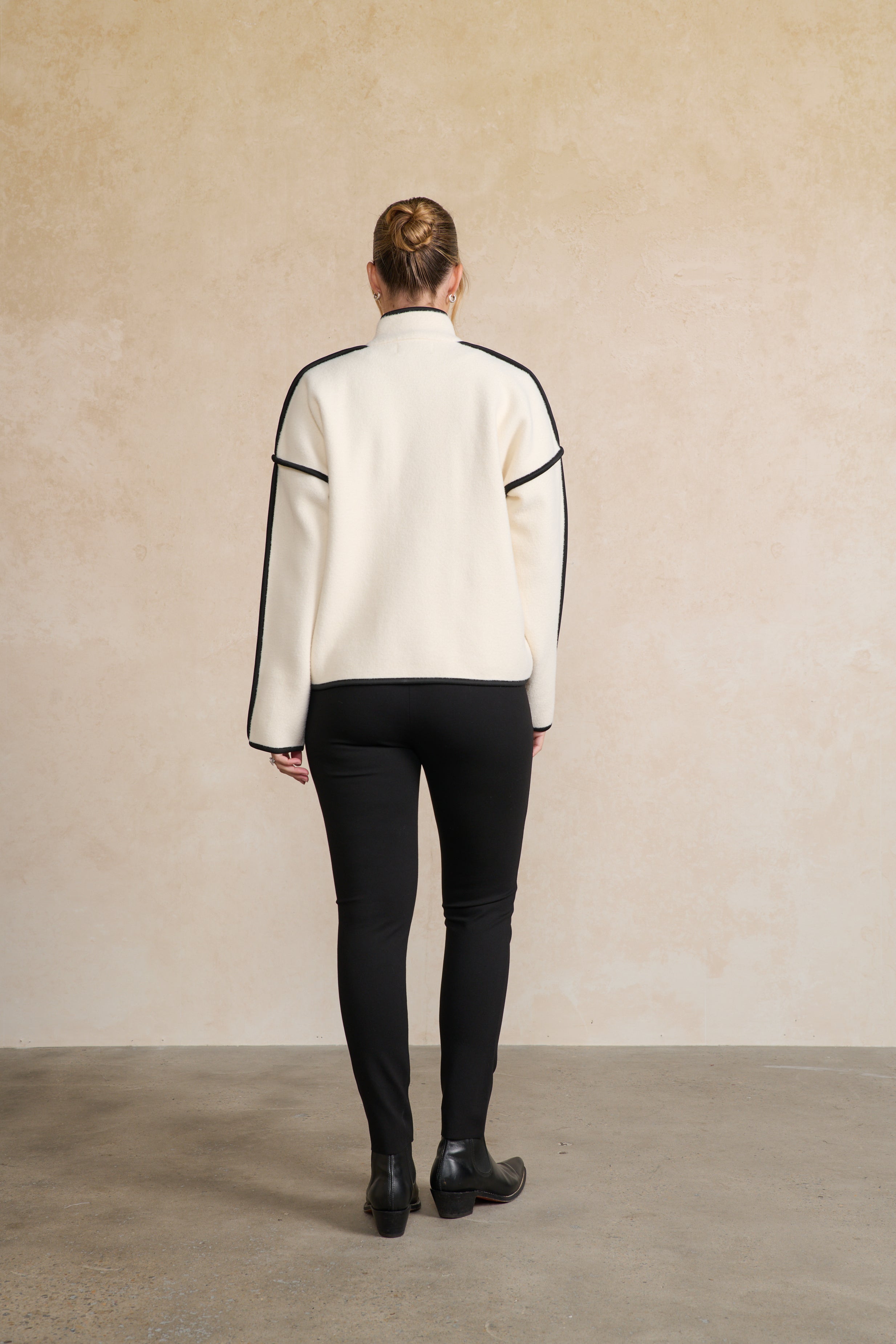 Person wearing a white sweater with black accents and black pants against a beige wall back angle