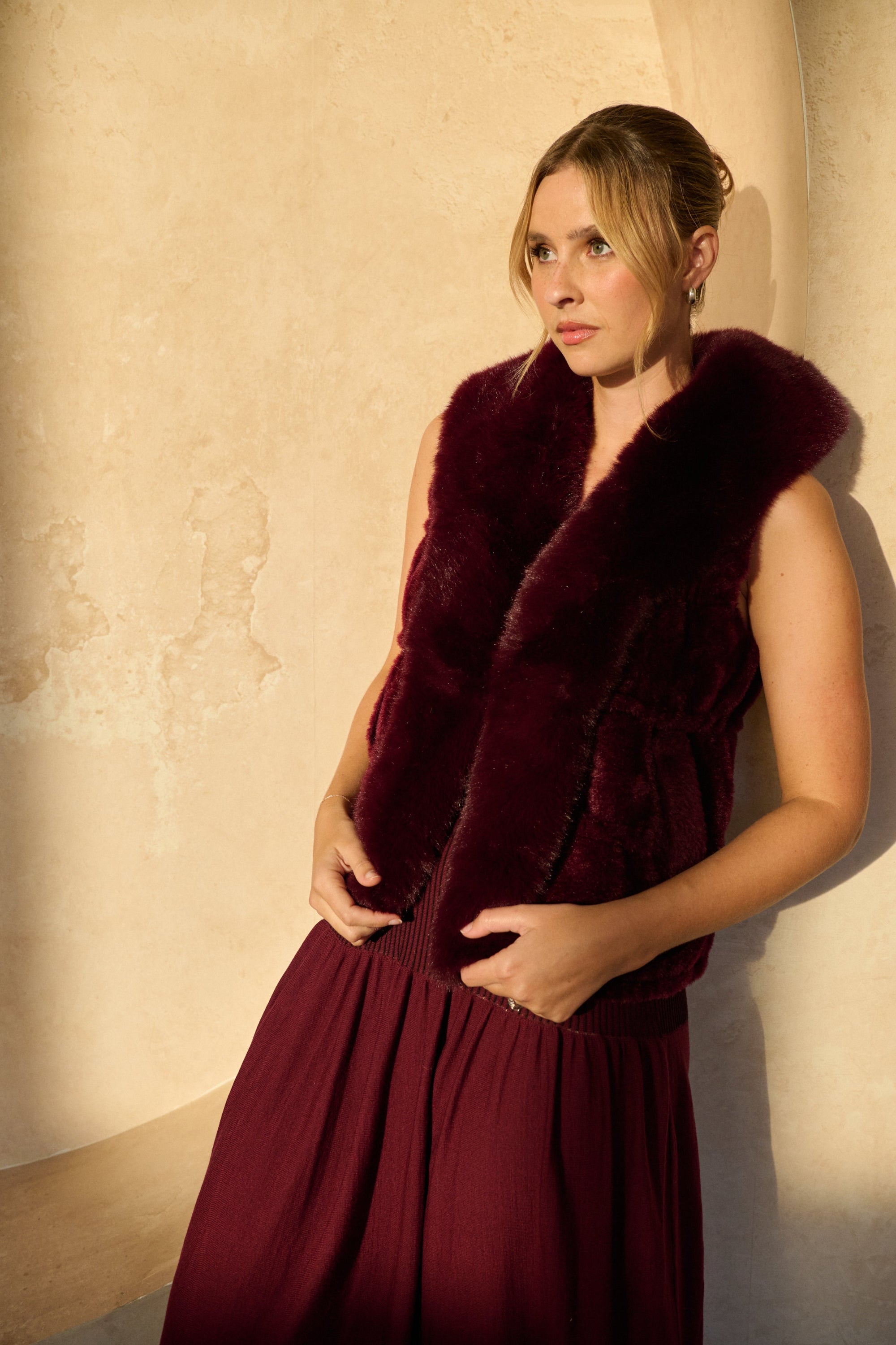 Woman wearing a burgundy fur vest over a dress against a beige wall.