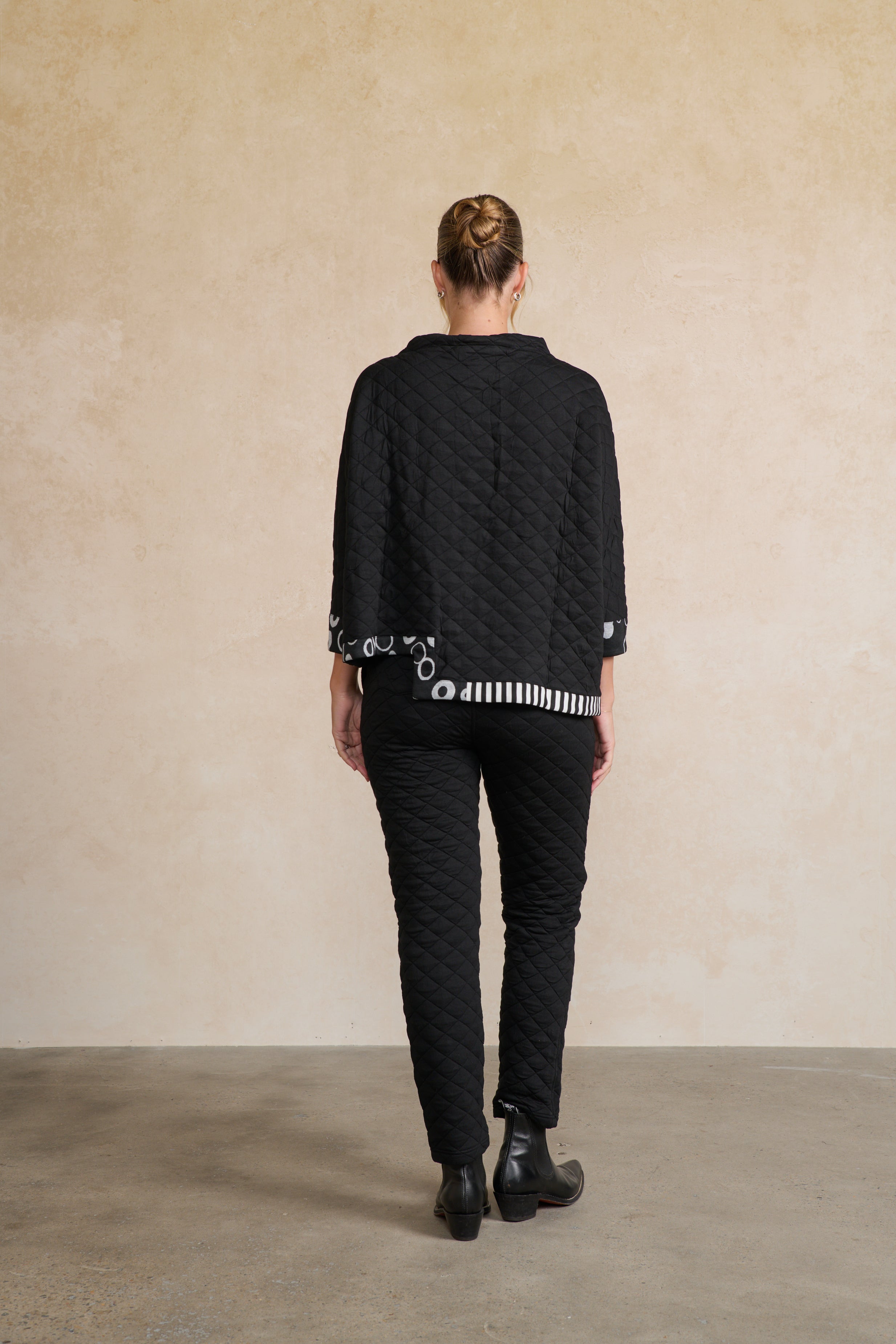 Person wearing a black quilted jacket and pants against a beige wall back angle