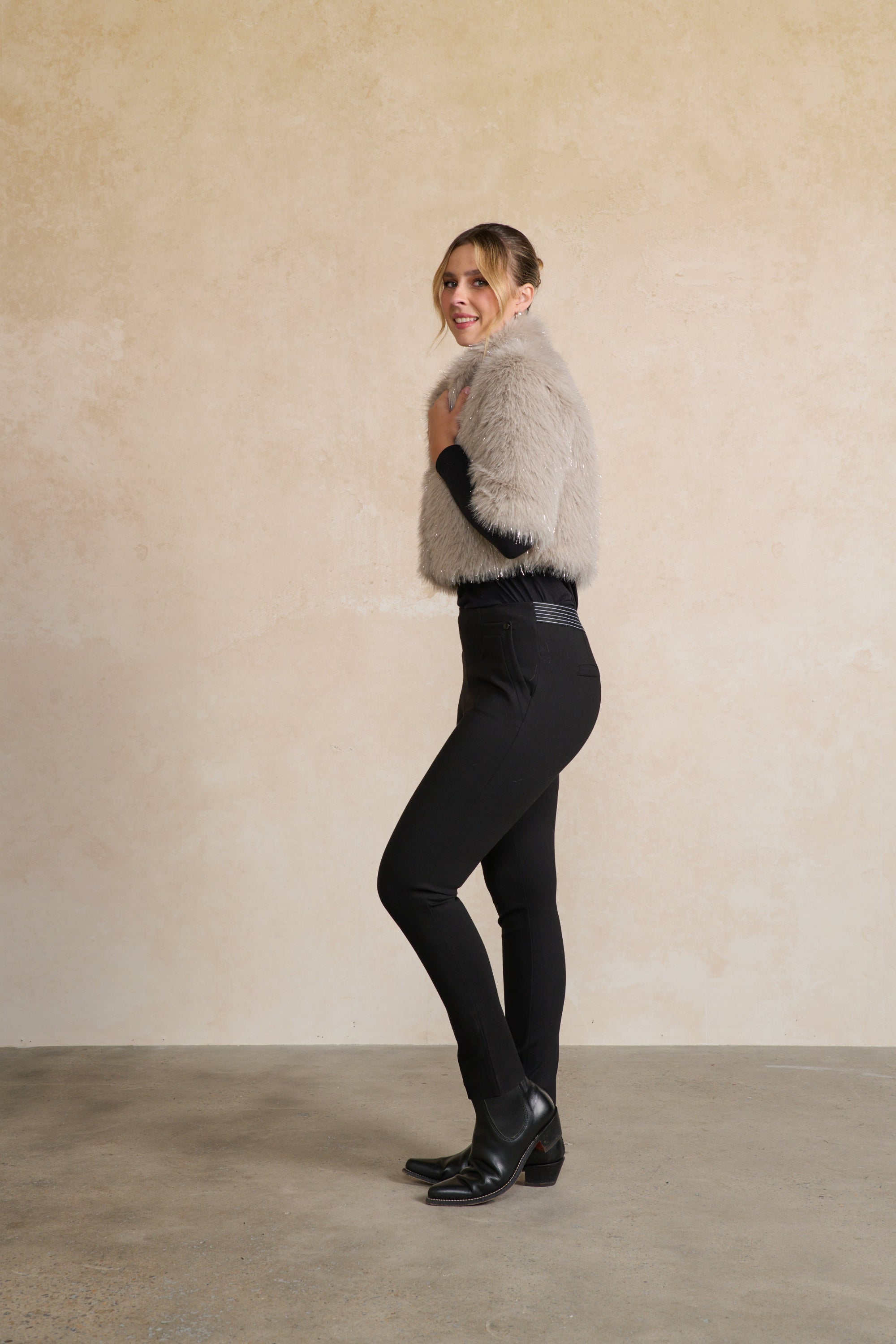 Person wearing a gray fur vest over a black outfit against a beige wall.