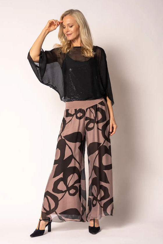 A woman modeling a sheer black mesh top with wide flutter sleeves and matching wide-leg pants featuring a black floral pattern on a dusty mauve background, paired with black heels, against a plain light background.