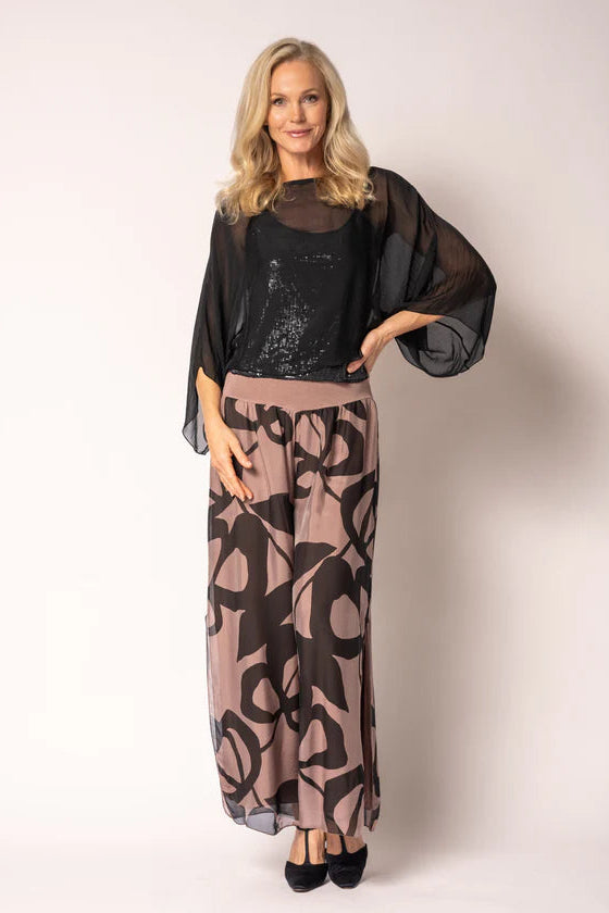 A woman modeling a two-piece outfit consisting of a sheer black long-sleeve top over a black sequined camisole, paired with a high-waisted maxi skirt in dusty pink and black abstract pattern, on a light beige studio background.
