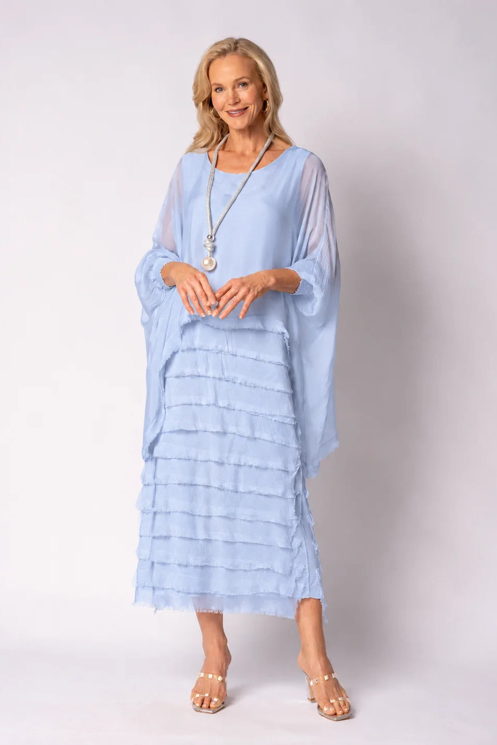 A woman modeling a light blue tiered maxi dress with sheer draped sleeves and a long silver pendant necklace, standing against a light gray studio background.