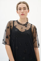 Woman wearing a black dress with sheer sleeves and floral embellishments on a white background