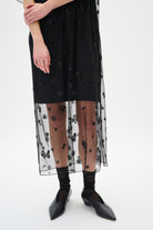 Black dress with floral embroidery, sheer overlay, black tights, and black shoes on a white background