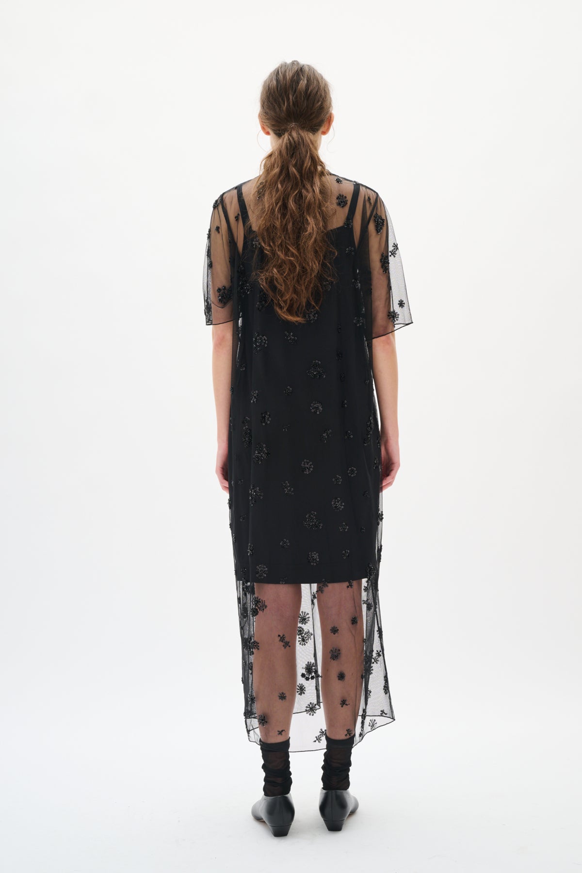 Woman wearing a black dress with sheer overlay and floral patterns on a white background back angle