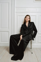 Woman wearing a black suit sitting against a white paneled wall.