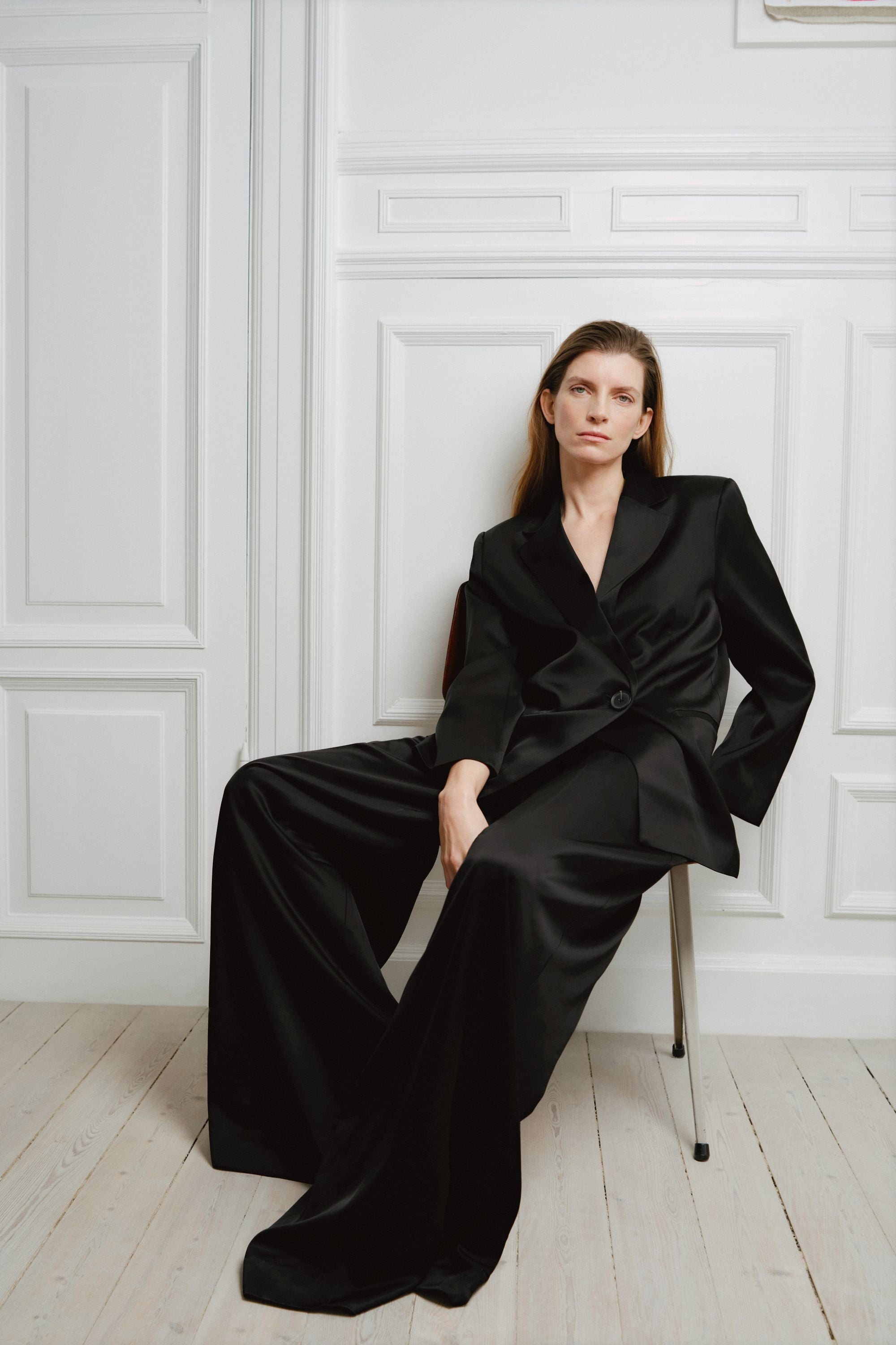 Woman wearing a black suit sitting against a white paneled wall.