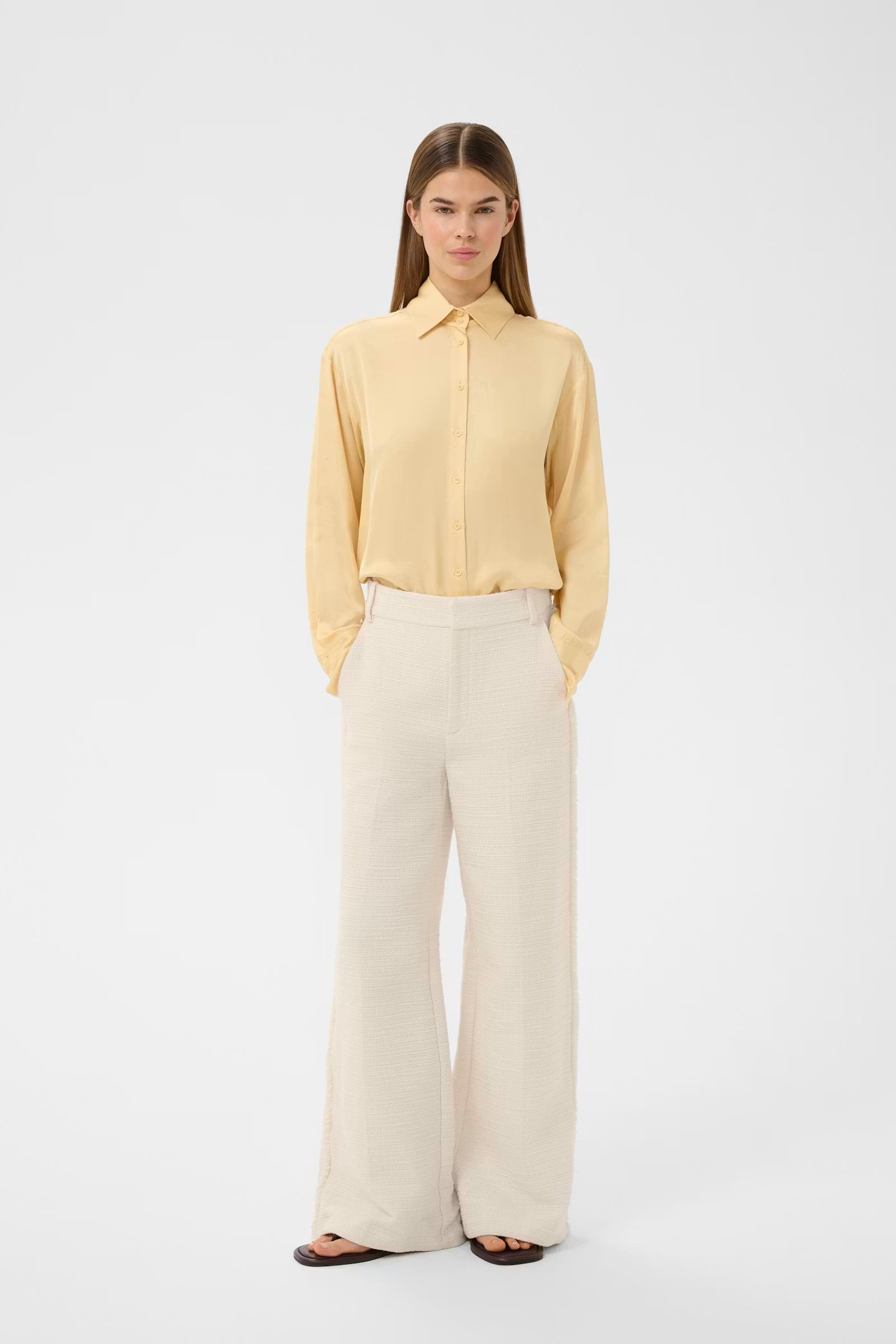 A woman modeling a pale yellow long-sleeve button-up shirt paired with wide-leg cream trousers and dark sandals, standing against a plain light gray studio background.