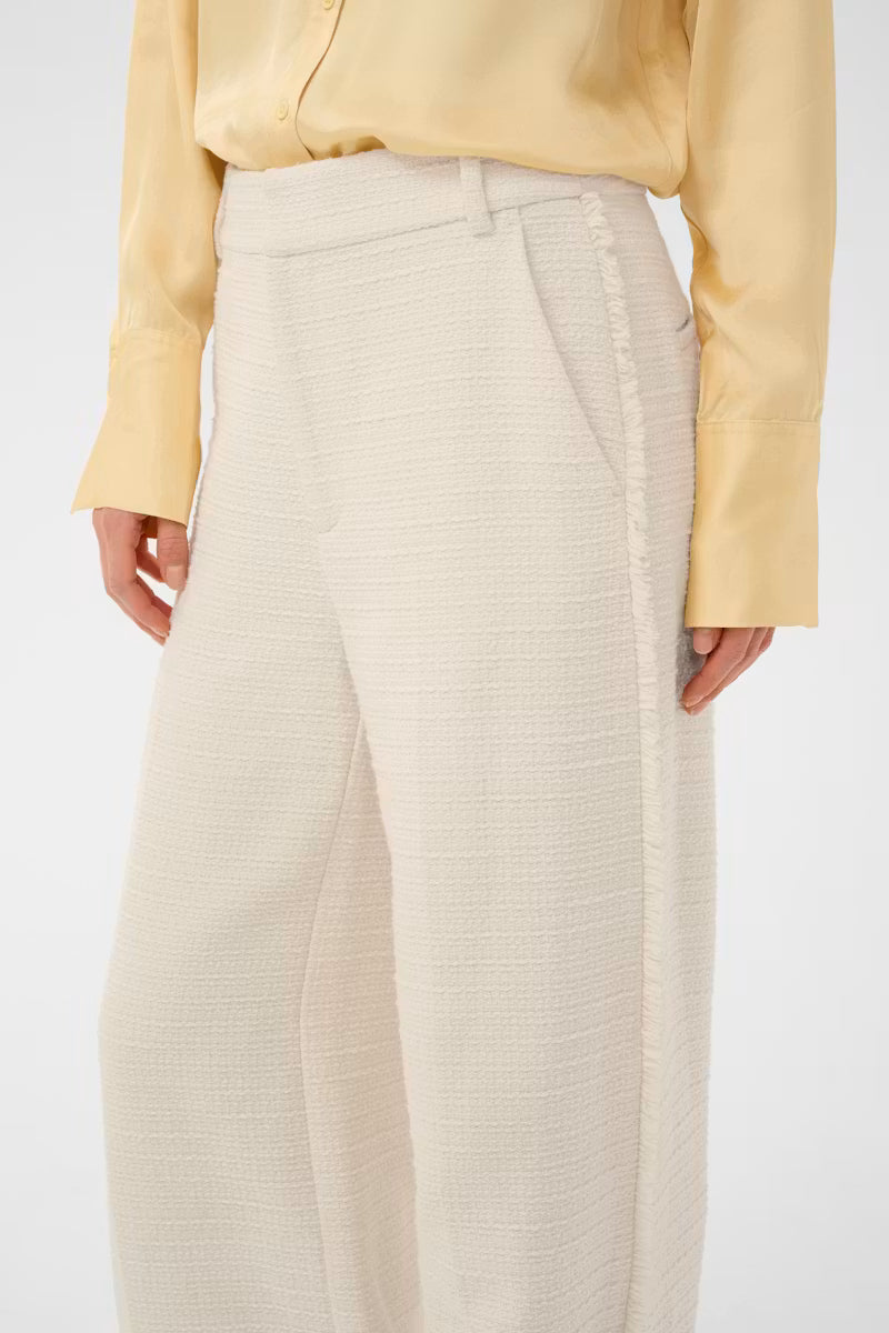 A close-up of a model wearing high-waisted, wide-leg trousers in a textured cream fabric, paired with a pale yellow satin blouse against a plain white background.