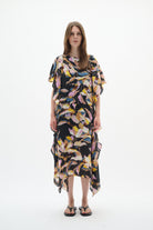 A woman modeling a loose, short-sleeved kaftan dress with a black base and a colorful abstract leaf print in pink, yellow, blue, and white, set against a plain white studio background.