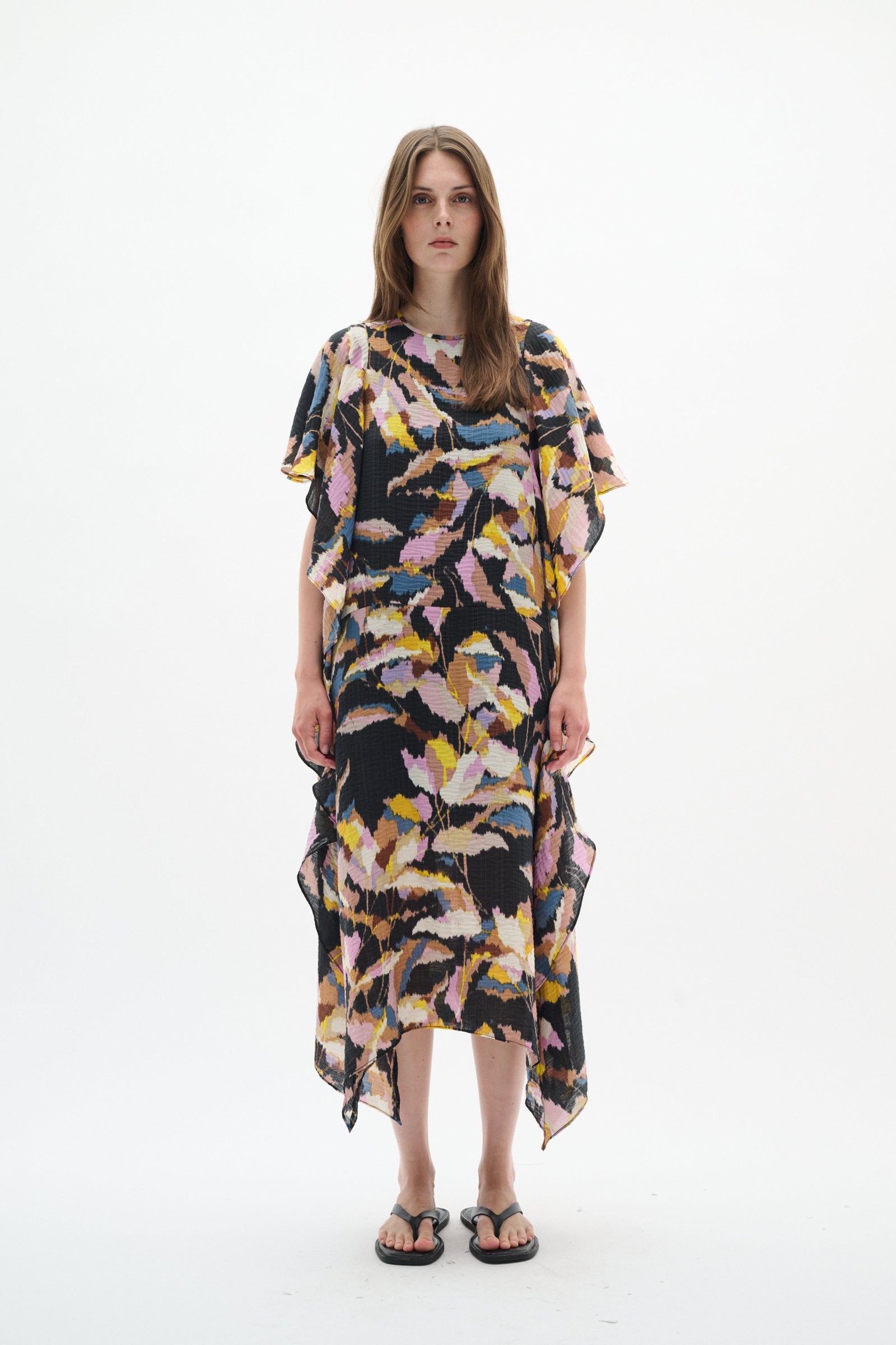 A woman modeling a loose, short-sleeved kaftan dress with a black base and a colorful abstract leaf print in pink, yellow, blue, and white, set against a plain white studio background.