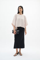 A woman modeling a light blush pink draped short-sleeve top and a black midi skirt, holding a black clutch, against a plain white studio background.