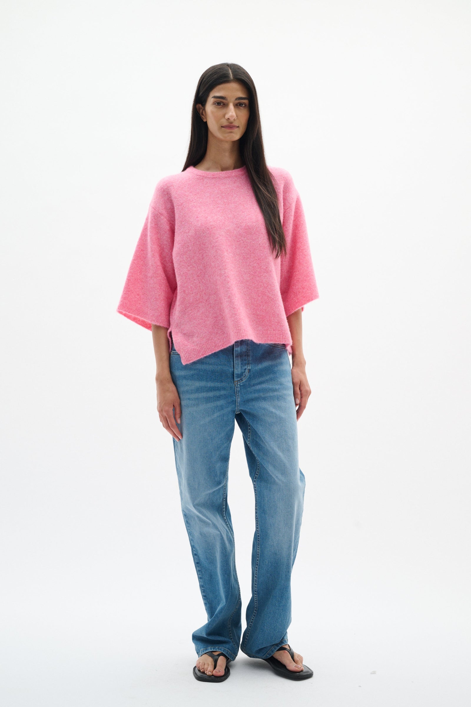 A woman modeling a pink short-sleeve knit top with blue jeans and black sandals, standing against a plain white studio background.