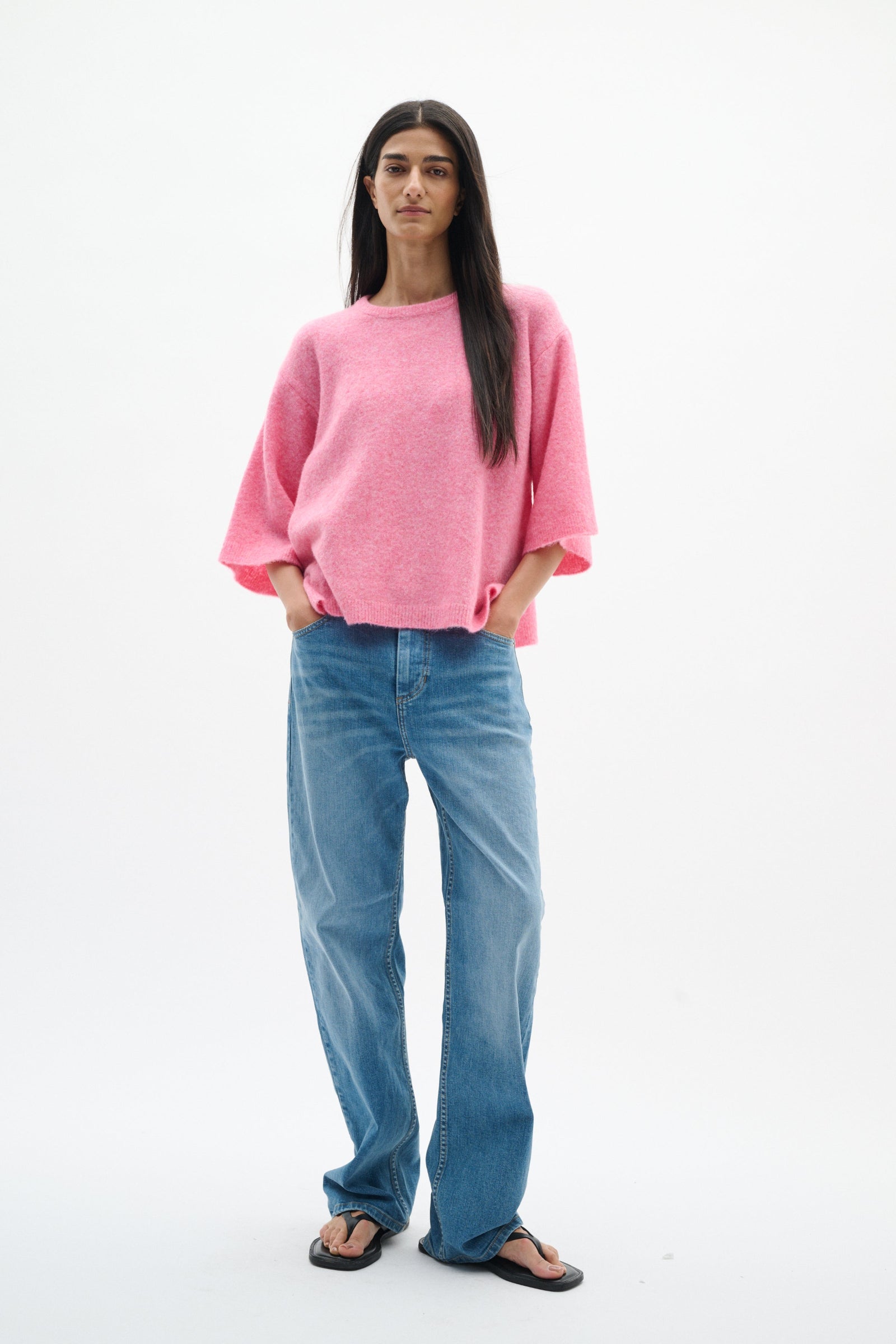 A woman modeling a pink knit sweater and blue jeans against a plain white studio background.
