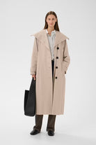 A woman stands against a light gray studio background wearing a long beige trench coat over a white shirt and dark pants, while holding a large black tote bag.