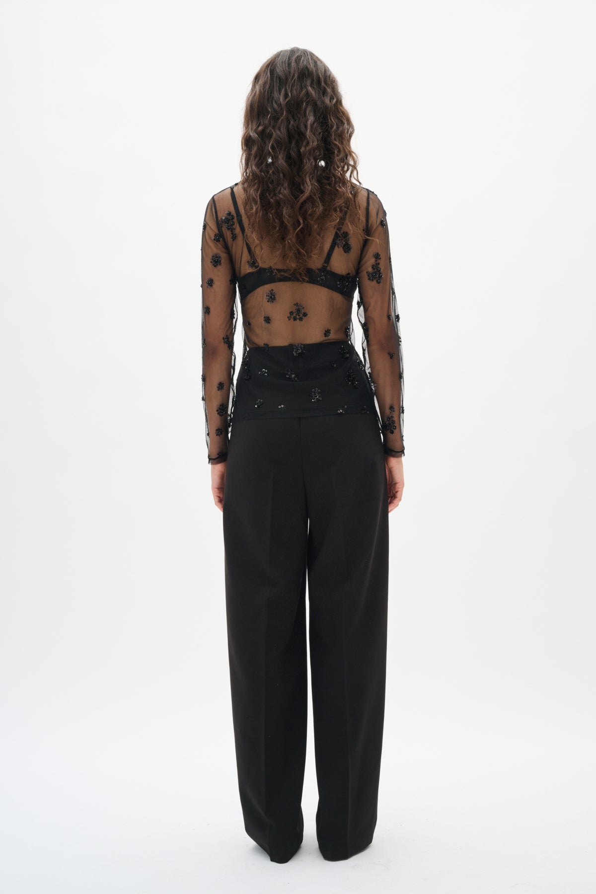 Person wearing a sheer top with black polka dots and high-waisted black pants on a white background back angle