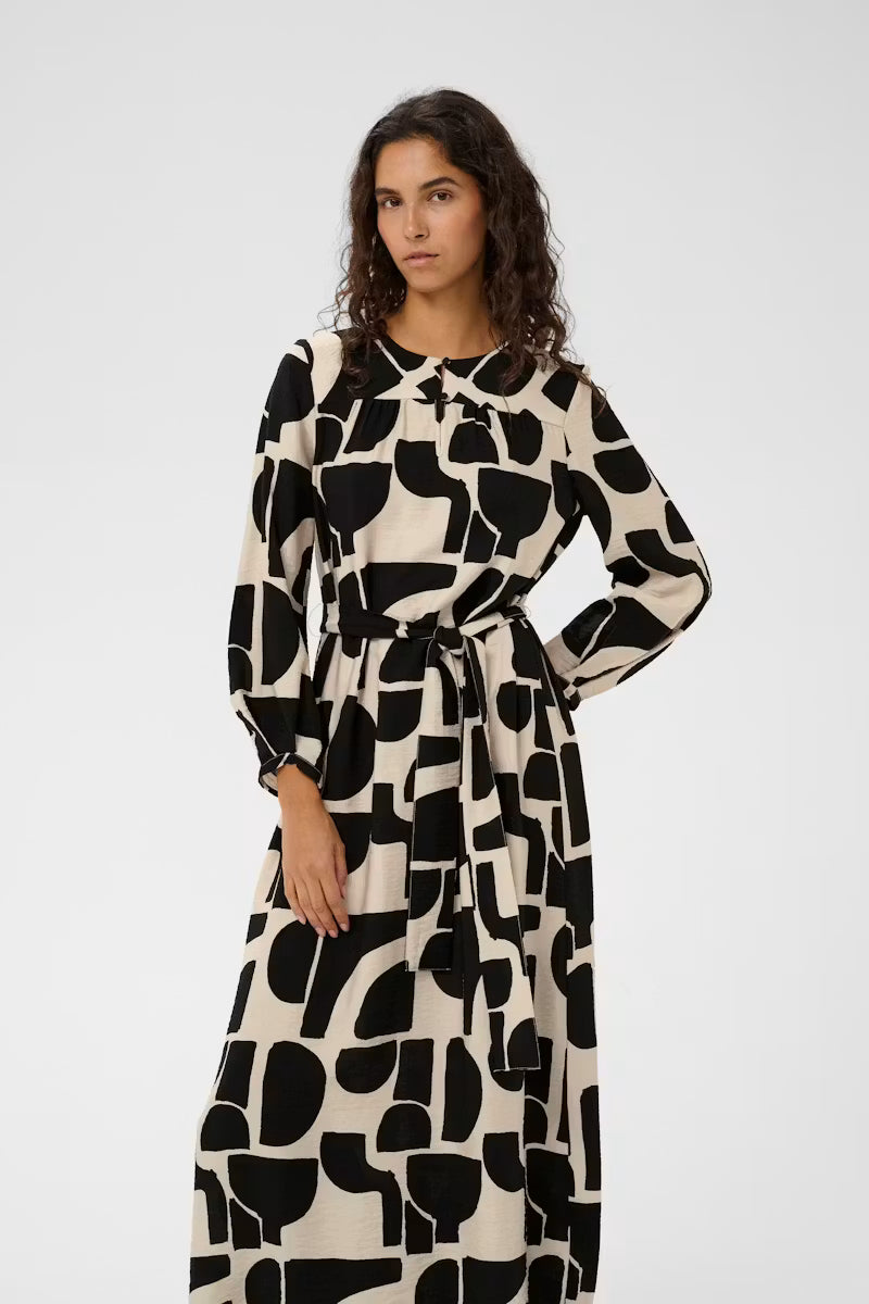 A woman modeling a long-sleeved midi dress with a black and cream abstract geometric pattern, featuring a collar and a matching tie belt at the waist, set against a plain light gray background.
