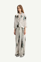 A woman modeling a light gray two-piece outfit with an abstract green and black watercolor print, consisting of a cropped jacket and matching wide-leg trousers, against a plain light gray background.