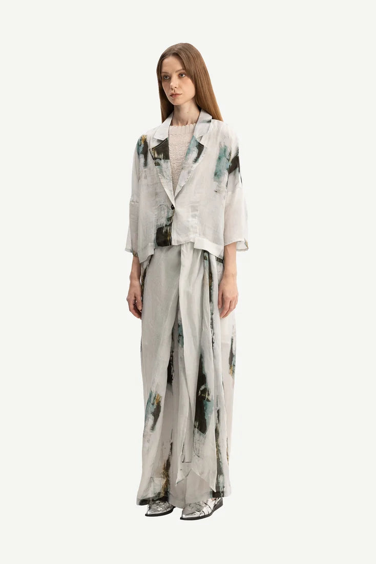 A woman modeling a light gray two-piece outfit with an abstract green and black watercolor print, consisting of a cropped jacket and matching wide-leg trousers, against a plain light gray background.