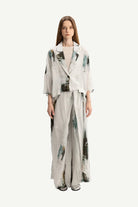 A model stands against a plain light gray studio background wearing a matching two-piece outfit consisting of a cropped, three-quarter sleeve jacket and wide-leg trousers, both in a light gray base with abstract watercolor-like green and black patterns.